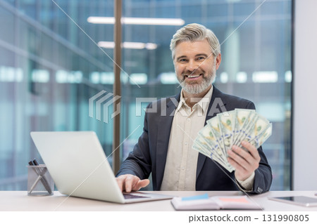 Successful middle-aged businessman smiling at the camera, holding a fan of dollar banknotes while actively working on a laptop in a modern office, representing financial success and remote income 131990805