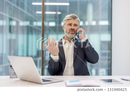 Mature businessman with grey hair and beard at office desk, stressed and gesturing while on an urgent phone call, conveying frustration and serious corporate concern 131990825