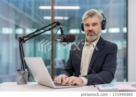 Mature man with headphones and a microphone broadcasting a podcast or online radio show, sitting at a desk and typing on a laptop, creating engaging content Mature man with headphones and a microphone broadcasting a podcast or online radio show, sitting at a desk and typing on a laptop, creating engaging content 131990860