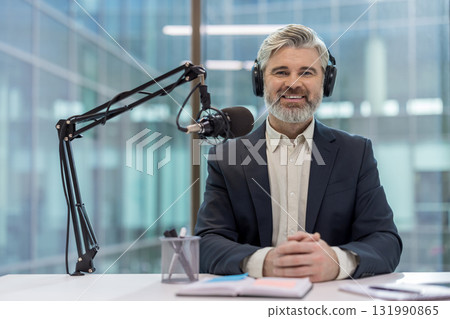 Smiling mature man wearing headphones and a suit jacket, sitting at a desk with a microphone and a notebook, broadcasting a podcast in a modern office with glass windows Smiling mature man wearing headphones and a suit jacket, sitting at a desk with a microphone and a notebook, broadcasting a podcast in a modern office with glass windows 131990865