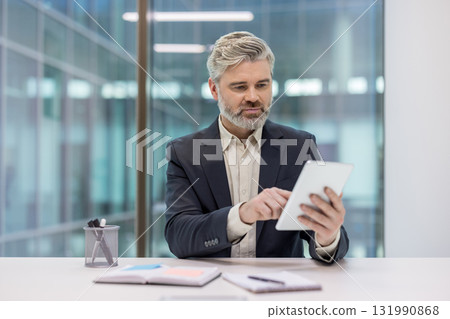 Businessman with a beard using a digital tablet at a modern office desk, interacting with technology while focusing on work tasks and corporate communication 131990868