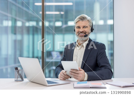 Mature businessman with gray hair and beard wearing a headset and holding a digital tablet, smiling confidently at his laptop while providing remote customer service in a modern office 131990898