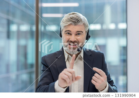 Mature man with a beard and headset talking, actively communicating with customers, providing support, and gesticulating with his finger in a modern office environment Mature man with a beard and headset talking, actively communicating with customers, providing support, and gesticulating with his finger in a modern office environment 131990904