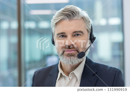 Mature businessman with gray beard wearing a headset and smiling, providing professional customer service and technical support from a modern office environment, confident and friendly 131990919
