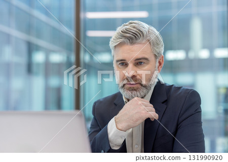Mature businessman having a pensive expression, strategizing in a modern office environment while looking at a laptop screen, conveying deep thought and decision-making for business success 131990920