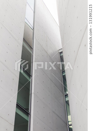 Tall concrete structures with large glass windows create a modern urban environment. The design showcases sharp lines and minimalistic features under a cloudy sky 131991215