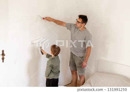 A father and his young son are busy painting the walls in a bedroom. The man is using a roller while the boy is helping with a small brush, creating a teamwork atmosphere 131991250