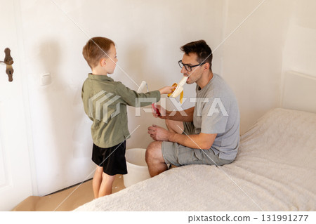 A father and his young son are painting the bedroom walls, applying a fresh coat of white paint. The boy is excitedly helping his dad while having fun in the neat, bright room 131991277