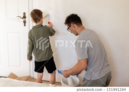 A man and his young son are whitewashing the walls of their bedroom. The boy uses a small roller while the father prepares paint, creating a bonding experience 131991284