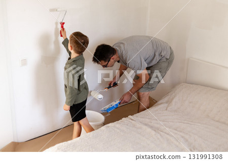 A man is whitewashing the walls in a bedroom while his young son assists him. Both enjoy a weekend activity that strengthens their bond and adds a fresh look to the room 131991308