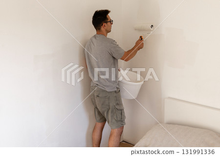 A man paints the walls of a bedroom with white paint using a roller. He stands focused, holding a paint bucket, creating a fresh, bright look in the room A man paints the walls of a bedroom with white paint using a roller. He stands focused, holding a paint bucket, creating a fresh, bright look in the room 131991336