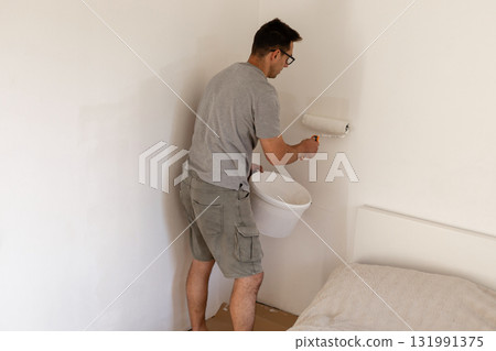 A man is busy painting the walls of a bedroom with a roller. He is using a bucket to hold the paint as he works on making the walls look fresh and clean 131991375