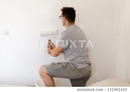 A man is in a bedroom applying a fresh coat of white paint to the walls using a roller. He stands on a stool for better reach, involved in home renovation work 131991420