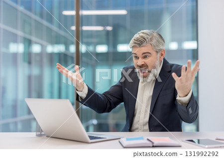 Agitated middle-aged businessman experiencing extreme stress and anger at his desk, reacting to a problem with his laptop in a modern office environment 131992292