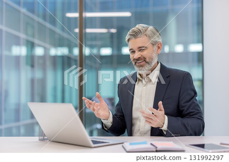 Smiling mature businessman having a video call on laptop displaying online communication and virtual meeting ,concept while gesturing during a remote discussion in a modern office Smiling mature businessman having a video call on laptop displaying online communication and virtual meeting ,concept while gesturing during a remote discussion in a modern office 131992297