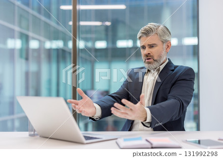 Stressed businessman in modern office experiencing confusion and frustration, gesturing at a laptop with a look of disbelief and concern over a technology problem 131992298