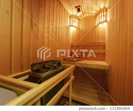 Modern empty wooden sauna room with warm lighting, stacked rocks on the heater, and wooden accessories. Perfect for wellness, spa, and relaxation concepts 131995069