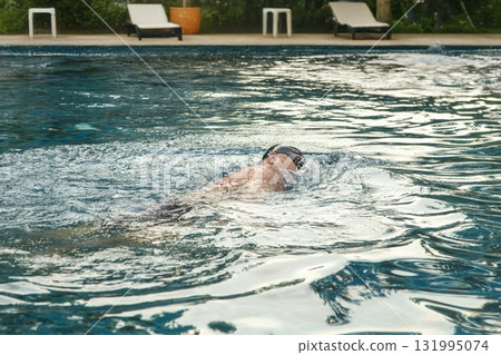 Man enjoying a relaxing, healthy swim in a beautiful pool on a sunny day. A perfect, tranquil escape for wellness and self-care. 131995074