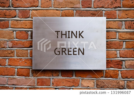 A silver sign displays the words THINK GREEN in bold, dark letters. The sign is affixed to a textured red brick wall outdoors during the daytime 131996087