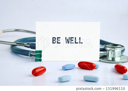 A Stethoscope is with several pills surrounding a card that reads BE WELL, on a white surface. A green clip holds the card up reminding us to stay healthy 131996110
