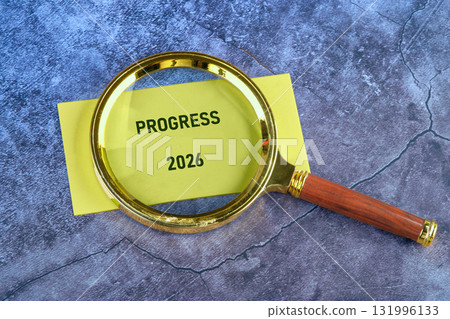 A yellow paper showing the word Progress and the year 2026 sits on a dark surface, as a gold and wood magnifying glass magnifies the potential for improvement in the future A yellow paper showing the word Progress and the year 2026 sits on a dark surface, as a gold and wood magnifying glass magnifies the potential for improvement in the future 131996133