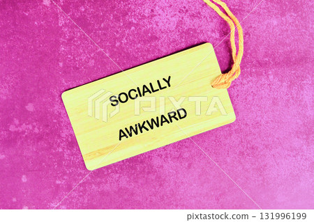 A wooden tag with the words SOCIALLY AWKWARD in block letters is placed on a textured, pink-purple background with a small yellow string to the side 131996199
