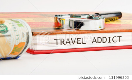A rolled stack of United States currency sits beside a Travel Addict book with a magnifying glass on top, suggesting a journey and the cost involved 131996203