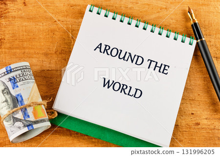 A notepad with the words AROUND THE WORLD sits next to a roll of one hundred dollar bills tied with twine and a black fountain pen on a wooden surface A notepad with the words AROUND THE WORLD sits next to a roll of one hundred dollar bills tied with twine and a black fountain pen on a wooden surface 131996205