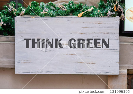 A whitewashed wooden sign displays the message THINK GREEN. Festive evergreen garland and pinecones adorn the top, promoting eco-consciousness in a natural setting A whitewashed wooden sign displays the message THINK GREEN. Festive evergreen garland and pinecones adorn the top, promoting eco-consciousness in a natural setting 131996305