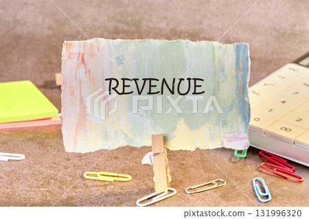 A colorful note with the word REVENUE is displayed with a wooden clothespin. Nearby are paperclips, sticky notes, and a calculator on a textured surface A colorful note with the word REVENUE is displayed with a wooden clothespin. Nearby are paperclips, sticky notes, and a calculator on a textured surface 131996320