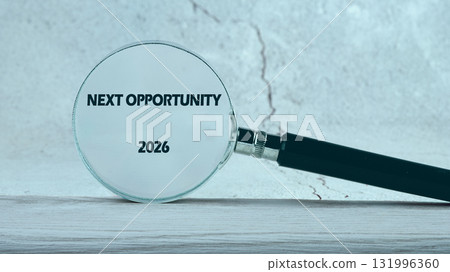 A magnifying glass focuses on the phrase NEXT OPPORTUNITY and the number 2026, displayed against a mottled gray background, resting on a textured light wood surface 131996360