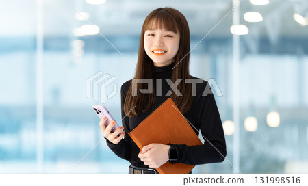 Smiling female businesswoman holding a smartphone and a binder 131998516