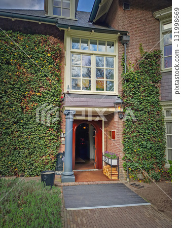 Charming, traditional brick building entrance with facade covered in climbing ivy, framing a prominent bay window and a red, wooden doorway with a stack of firewood beside adds to the rustic aesthetic Charming, traditional brick building entrance with facade covered in climbing ivy, framing a prominent bay window and a red, wooden doorway with a stack of firewood beside adds to the rustic aesthetic 131998669