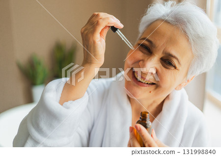 Woman applying natural drops cosmetics oil to face skin. Mature adult old woman applying nourishing anti age serum with pipette doing daily beauty routine. Senior woman gets kin scare facial treatment 131998840