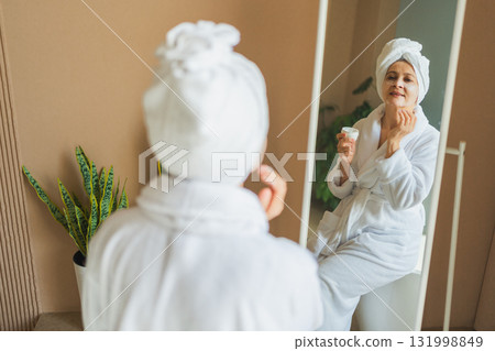 Mature adult old woman looking at mirror applying nourishing anti age face cream doing daily beauty routine. Senior woman with white mask creme on skin gets facial treatment Skin care aging skincare 131998849