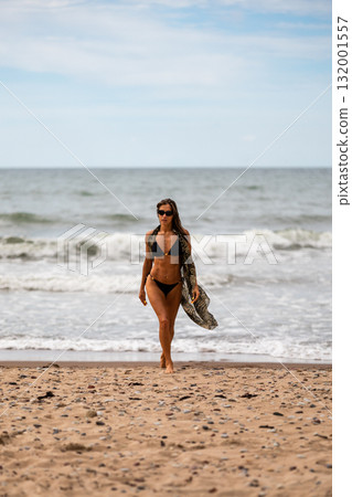 A joyful woman strolls along the beach, embracing the sun and waves in her stylish bikini while enjoying nature 132001557
