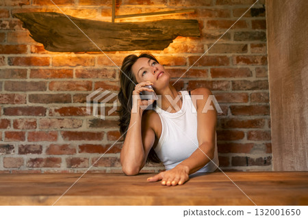 A woman in a white tank top thoughtfully engaged in a phone conversation in a warm, rustic environment. A woman in a white tank top thoughtfully engaged in a phone conversation in a warm, rustic environment. 132001650