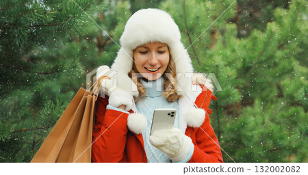 Happy smiling woman with phone, shopping bag in winter against Christmas tree, girl using smartphone 132002082