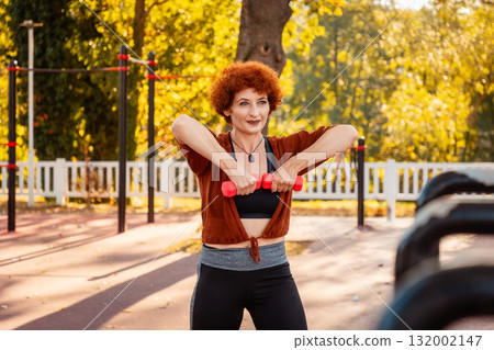 Fitness. Smiling adult pretty Caucasian ginger hair woman training in sports playground. Sunny autumn park in background. Athlete's hands lift up dumbbells Fitness. Smiling adult pretty Caucasian ginger hair woman training in sports playground. Sunny autumn park in background. Athlete's hands lift up dumbbells 132002147