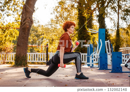 Side view of Caucasian pretty adult woman wearing sports clothes stretching using dumbbells. Fitness in city park Side view of Caucasian pretty adult woman wearing sports clothes stretching using dumbbells. Fitness in city park 132002148