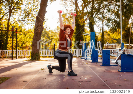 Adult Caucasian woman wearing sport clothes training in city park. Athlete does squats with dumbbells in her hands Adult Caucasian woman wearing sport clothes training in city park. Athlete does squats with dumbbells in her hands 132002149