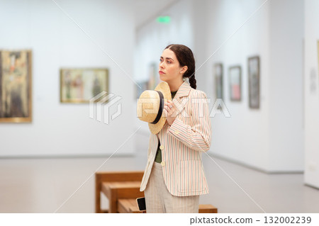 Mid shot of a young Caucasian woman in elegant jacket and trousers holds straw hat and posing in the art gallery. Painting in background. Concept of cultural education and Museum Day Mid shot of a young Caucasian woman in elegant jacket and trousers holds straw hat and posing in the art gallery. Painting in background. Concept of cultural education and Museum Day 132002239