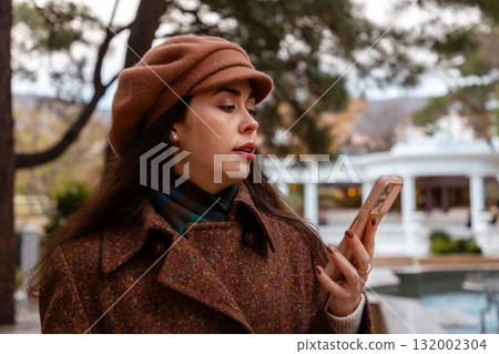 A stylish Caucasian woman walks through the autumn city while using her smartphone, surrounded by architecture and warm natural tones. 132002304