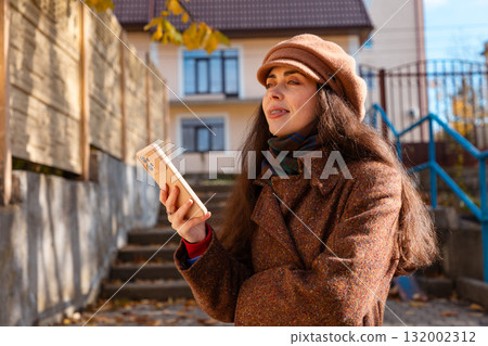 Portrait of young Caucasian woman smiling in a coat and hat uses the phone while standing on the steps. Lifestyle and modern city life 132002312