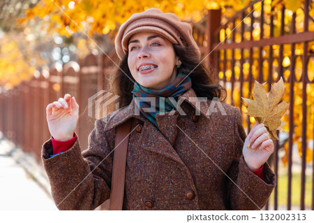 Portrait of young pretty Caucasian happy woman with braces on her teeth in a coat and hat holds bunch of autumn yellow leaves and waving hands, standing on the street. Lifestyle and urban life 132002313