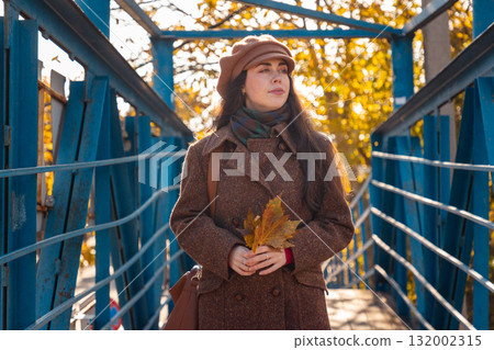 Mid shot of young pretty elegant Caucasian woman in a coat holds bunch of yellow leaves and walking on the bridge in city street on autumn day. Lifestyle and city life 132002315