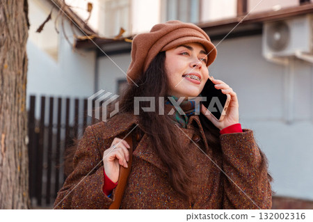 Portrait of young pretty Caucasian happy woman with braces on her teeth, wearing a coat and hat call with smartphone standing on the street and smiling. Mid shot. Lifestyle and urban life 132002316