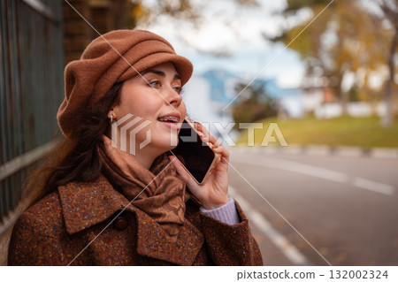 Side view of young beautiful happy Caucasian woman in a coat and hat calling taxi on phone in city on autumn day. Road in background. Lifestyle and city life 132002324