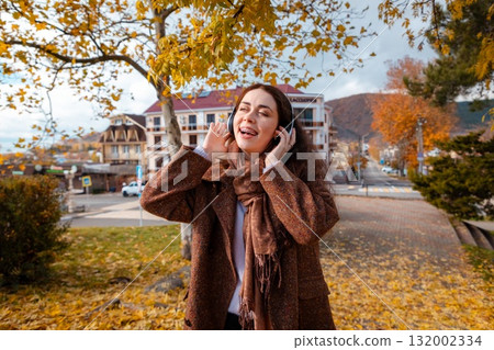 Mid shot of a happy young Caucasian pretty woman in a coat listening to music with headphones and singing in park. Concept of happiness 132002334