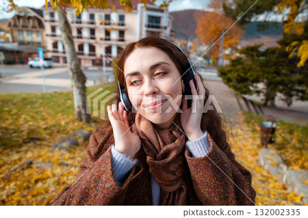 Close up portrait of a happy young Caucasian pretty woman in a coat listening to music with headphones in park. Concept of happiness Close up portrait of a happy young Caucasian pretty woman in a coat listening to music with headphones in park. Concept of happiness 132002335